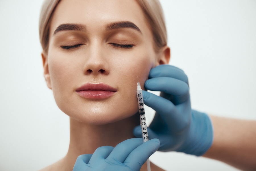 Wanna be perfect. Portrait of young pretty woman keeping eyes closed while doctors hand in blue medical gloves making an injection in her face