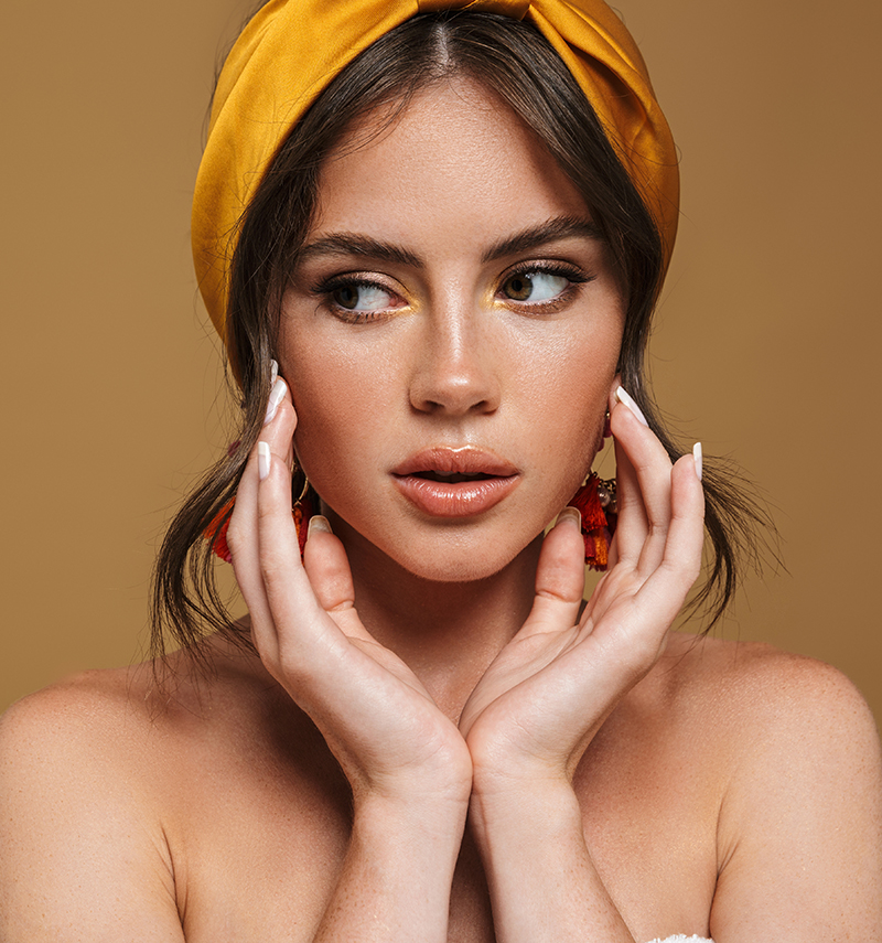 Close-up of a woman's face, wearing a yellow headband and earrings. She has her hands gently touching her face